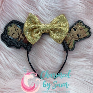 May include: A pair of black mouse ears with a gold glitter bow and two felt Groot characters. The characters are brown and green with a black outline. The text "Charmed by Sam" is visible below the ears.