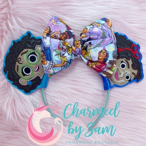 May include: A pair of blue and brown character headbands with a large bow featuring a colorful print of characters from the Disney movie Encanto. The bow is tied with a red ribbon. The headband is decorated with glitter. The text "Charmed by Sam" is visible below the headband.