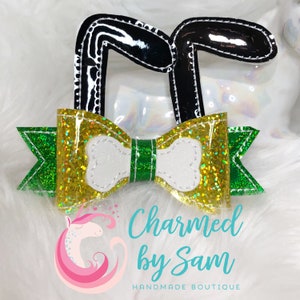 May include: A green and gold glitter bow with a white bone-shaped center, topped with black bunny ears. The bow is attached to a white fuzzy surface. The text "Charmed by Sam" and "HANDMADE BOUTIQUE" is below the bow.