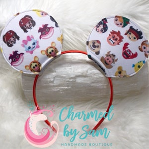May include: Red headband with white fabric mouse ears featuring a variety of cartoon character faces. The ears are decorated with characters from Disney movies like The Lion King, Aladdin, and The Little Mermaid. The headband has the text "Charmed by Sam" and "Handmade Boutique" printed on it.