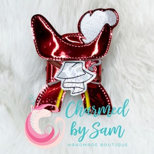 May include: Red and silver fabric pirate hat with a bell charm. The hat is made of shiny fabric and has a white fur trim. The charm is a silver bell with a white fabric bow. The hat is on a white fuzzy surface. "Charmed by Sam" is written in blue text below the hat.