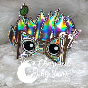 May include: A holographic bow with black embroidered eyes and a green leaf accent. The bow is surrounded by a flame-like design with a metallic sheen. The text "Charmed by Sam Handmade Boutique" is visible.