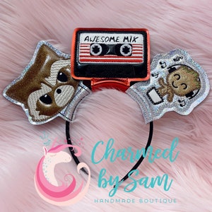May include: A headband with a silver glitter background featuring a cartoon image of Rocket Raccoon, a cassette tape with the text "Awesome Mix" and a cartoon image of Groot wearing headphones. The headband is black and has a pink and white unicorn logo with the text "Charmed by Sam Handmade Boutique".