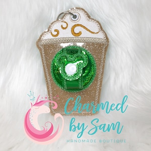 May include: A sparkly brown and white coffee cup keychain with a green lid and a white button. The keychain is decorated with a swirl of whipped cream and a gold design. The text "Charmed by Sam" and "HANDMADE BOUTIQUE" is visible below the keychain.