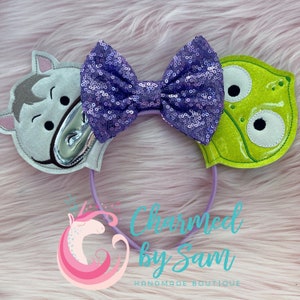 May include: Purple sequin bow headband with a green alien and a gray horse character on each side. The headband is purple and has the text "Charmed by Sam Handmade Boutique" on the bottom.