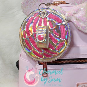 May include: A pink and iridescent keychain accessory with a circular design and a smaller, matching strap. The circular piece has a pink embroidered pattern and is outlined with silver rhinestones. The strap has a pink button. The text "Charmed by Sam" is visible.