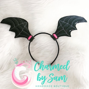 May include: Black felt bat wings headband with pink accents. The headband is on a white fuzzy surface. "Charmed by Sam" is written in teal text below the headband.