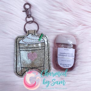 May include: A glittery gold coffee cup keychain with a pink heart and green straw, next to a bottle of "raspberries & whipped vanilla" hand sanitizer. The keychain has a silver clasp. The hand sanitizer is 1 fl oz / 29 ml.