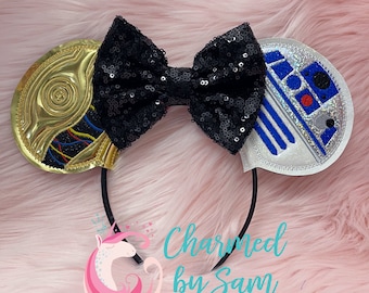 May include: A pair of black sequin Minnie Mouse ears with a gold and black Star Wars character on one ear and a blue and white Star Wars character on the other ear. The ears are on a pink fuzzy background.