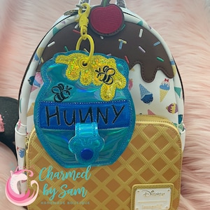 May include: A blue and yellow honey pot-shaped bag charm with a bee and the word "Hunny" embroidered on it. The charm is attached to a white and brown ice cream cone-themed backpack with a cherry on top.