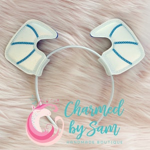 May include: White headband with two blue and white basketball-shaped ears. The headband is on a pink fuzzy surface. The text "Charmed by Sam" is below the headband. The text "HANDMADE BOUTIQUE" is below the text "Charmed by Sam".