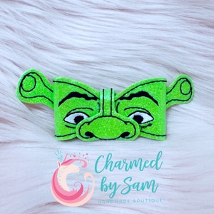 May include: Green glitter bow tie with a black outline of Shrek's face. The bow tie has a green glitter background with black outlines of Shrek's face, including his ears, eyebrows, eyes, nose, and mouth. The bow tie is on a white fuzzy background.