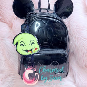 May include: Black Mickey Mouse-shaped backpack with attached ears and a green Oogie Boogie keychain. The keychain features a smiling Oogie Boogie with a colorful snake. The backpack has a glossy finish and a front zipper.