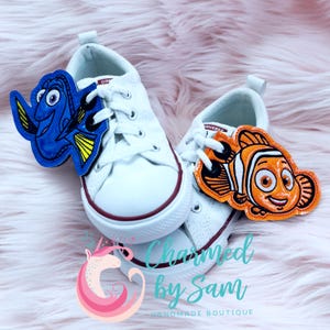 May include: A pair of white sneakers with blue and orange character shoe charms. The charms are of Dory and Nemo from the movie Finding Nemo. The charms are attached to the laces of the sneakers.