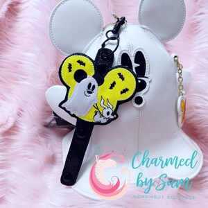 May include: A black and yellow Halloween-themed Mickey Mouse ear keychain with a ghost and a dog character. The keychain is attached to a black strap with a snap closure. "Charmed by Sam Handmade Boutique" is printed on the white background.