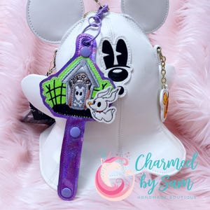 May include: White faux leather Mickey Mouse ears with a purple iridescent strap and a charm featuring a green and white haunted house with a ghost and a black and white Mickey Mouse head.