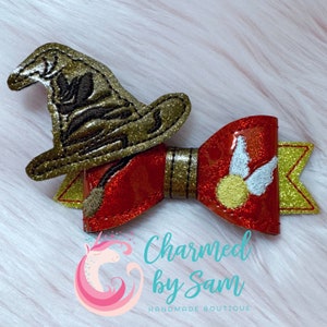 May include: A red and gold glittery hair bow with a gold witch hat and a golden snitch. The bow has a golden broom and a golden banner. The text "Charmed by Sam Handmade Boutique" is visible.