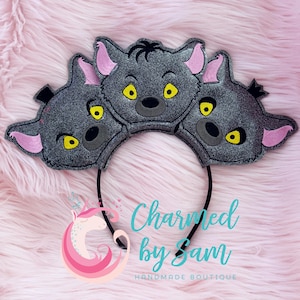 May include: Three-headed gray and black hyena ears with pink ears and yellow eyes on a black headband. The headband is decorated with a white unicorn logo with pink and blue accents and the text "Charmed by Sam Handmade Boutique".