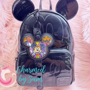 May include: Black Mickey Mouse-shaped backpack with matching ears. A Halloween-themed Mickey Mouse keychain is attached. The keychain features a colorful embroidered design of Mickey Mouse in a Halloween costume. The text "Charmed by Sam" is visible.