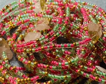 Op de afbeelding: Een opgerolde streng kralenjuwelen met afwisselend kleine roze, lichtgroene en metallic gouden glaskralen. De streng is geaccentueerd met verschillende onregelmatige, doorschijnende, lichtgele en heldere steensplinters verspreid over de lengte.