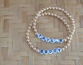 May include: Two gold beaded bracelets with the names "Brody" and "Taylor" spelled out in white beads.
