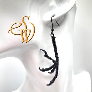 May include: A single black earring in the shape of a bird's claw. The claw has a textured surface and sharp talons. The earring hangs from a black hook. The background is a blurred white.