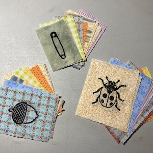 May include: Three piles of fabric squares with embroidered designs. One square shows a black safety pin, another a ladybird, and the third an acorn. The squares have scalloped edges and are in various floral and patterned fabrics.