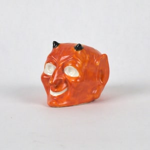 May include: A ceramic figurine of a red devil's head with black horns and white eyes. The devil is smiling.