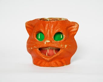 May include: An orange cat-shaped container with a textured surface, featuring bright green eyes and an open mouth revealing sharp white teeth. The object has pointed ears and a hollow top opening, displaying a weathered finish with visible paint wear.