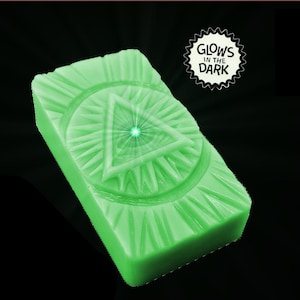 Puede incluir: Un objeto rectangular verde brillante con un diseño geométrico, que incluye un triángulo y líneas radiales. El objeto tiene una superficie texturizada y el texto "GLOWS IN THE DARK" es visible. Sobre un fondo negro.