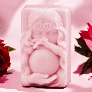 May include: A pink soap bar featuring a raised design of a woman holding a crystal ball. The woman has long hair and is wearing a headband. The soap is surrounded by pink roses.