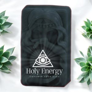 May include: Black soap bar with a white image of a woman's face and a triangle with an eye inside. The text "Holy Energy" and "Empower Your Aura" is printed below the image.