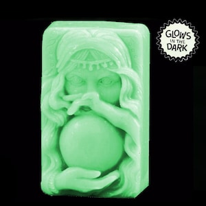 Puede incluir: Una pastilla de jabón verde con la cara de una mujer y un orbe brillante en sus manos. El jabón brilla en la oscuridad. "Glows in the Dark" está escrito en una pegatina en forma de estrella.
