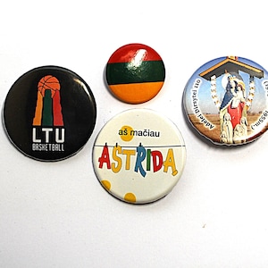 May include: Four round pins with various designs. One pin features the text "LTU BASKETBALL" with a basketball graphic. Another has the Lithuanian flag colors. A third pin says "ASTRIDA". The last pin has a religious image.