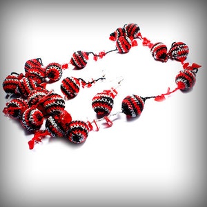 Può includere: Una collana di uncinetto rosso, nero e bianco con perline rotonde. Le perline sono a strisce rosse e bianche, e alcune hanno accenti neri.