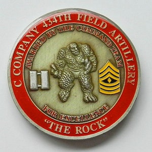 May include: A red, white, and gold coin with the words "Company 434th Field Artillery" and "Awarded by the Command Team" around the edge. The center of the coin features a cartoon-like image of a muscular figure lifting weights, with the words "For Excellence" and "The Rock" below.