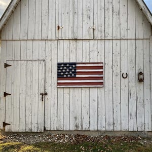 May include: A wooden American flag hangs on the weathered white exterior of a barn. The flag features a blue field with white stars and red and white stripes. A horseshoe and lantern are also mounted on the barn.