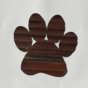Op de afbeelding: Een wanddecoratie in de vorm van een pootafdruk gemaakt van bruin golfplaatmetaal. Het object heeft een getextureerd, geribbeld oppervlak met een roestige uitstraling, drie driehoekige uitsparingen voor de tenen en een klein rond gat links.