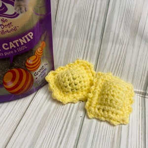 May include: Two yellow crocheted catnip toys shaped like ravioli. The toys are on a light wood surface. The text on the bag behind the toys reads "No Dogs Allowed!" and "Catnip".