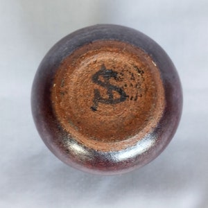 May include: The bottom of a dark brown ceramic vase with a silver glaze. The vase has a black "S" mark on the bottom.