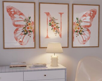 May include: Three framed wall art pieces. Two feature butterfly wing designs in coral and white, adorned with floral arrangements. The third displays the letter "N" with a floral design. The frames are a light brown color.