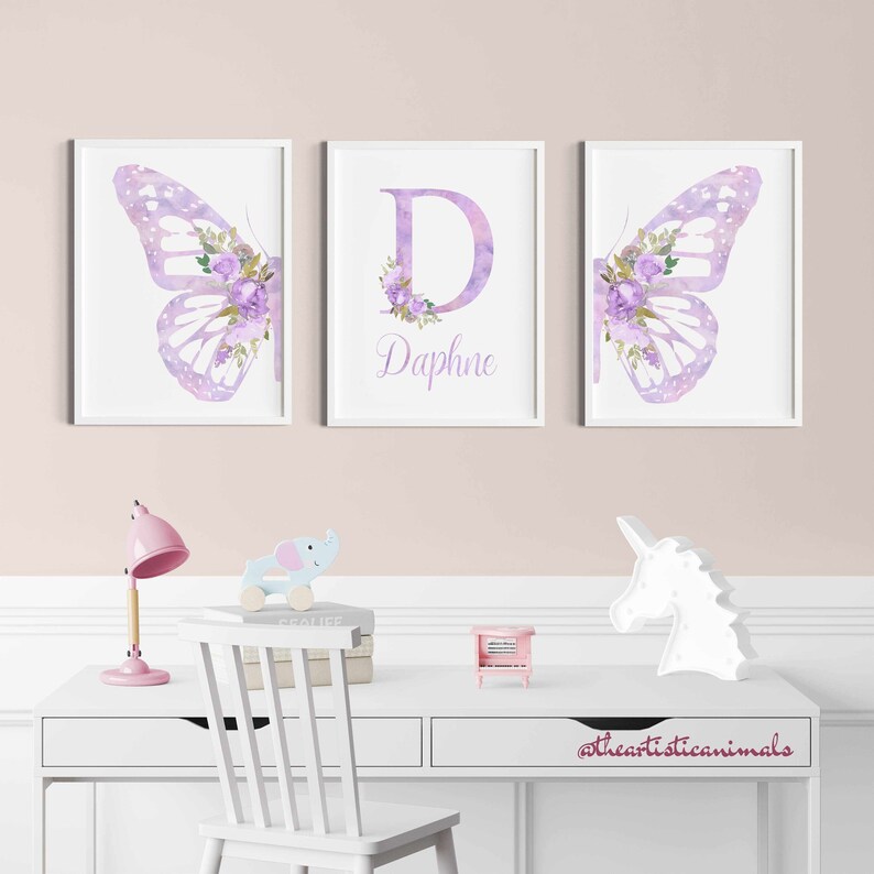 Op de afbeelding: Drie ingelijste prints met een aquarel vlinderontwerp. De middelste print heeft de letter "D" met de naam "Daphne" eronder. De prints zijn in een witte lijst met een witte achtergrond.