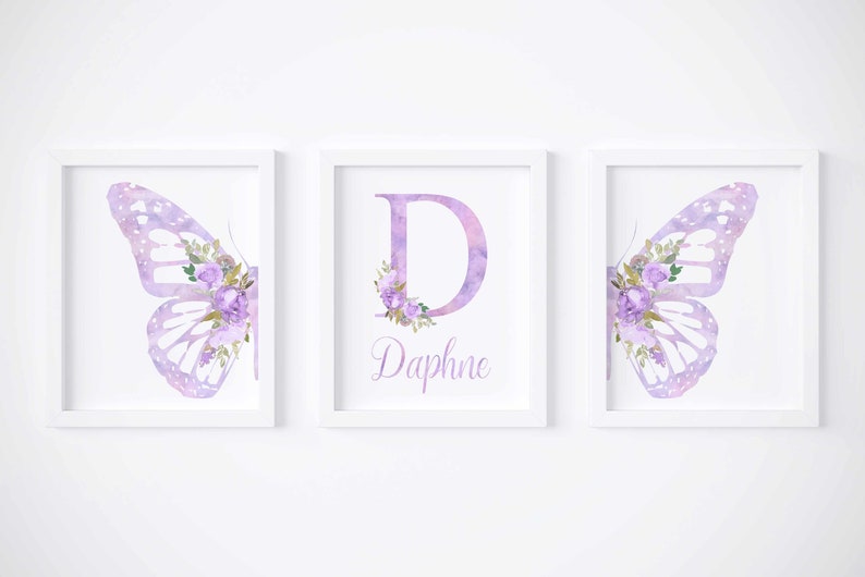 Op de afbeelding: Drie ingelijste prints met een aquarel vlinderontwerp. De middelste print heeft de letter "D" met paarse bloemen en de naam "Daphne" eronder. De prints zijn perfect voor een kinderkamer of babykamer.
