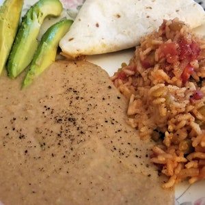 May include: A white plate with a serving of refried beans, rice, avocado slices, and a tortilla. The beans are topped with black pepper.