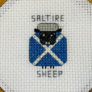 May include: A white cross-stitch embroidery hoop with a black sheep wearing a blue and white saltire pattern. The text "Saltire Sheep" is stitched above and below the sheep.