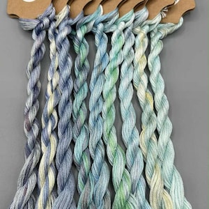 May include: A selection of hand-dyed embroidery floss in various shades of blue, green, and cream, labelled 'Light Fantastic', set 3 of 4. Each skein is twisted and attached to a brown card with the 'Midge Porter Design' logo. Suitable for crafts.