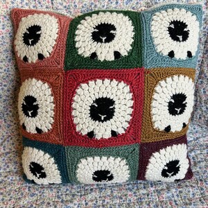 Peut inclure: Un coussin crocheté avec un motif patchwork composé de neuf carrés. Chaque carré présente un mouton blanc avec des yeux noirs et un nez noir au centre. Les carrés sont dans des tons de rouge, vert, bleu et marron.
