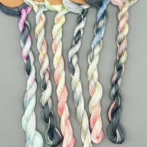 May include: A collection of hand-dyed embroidery floss in a variety of colors, including pink, blue, green, and gray. Each skein is labeled with a brown tag. The text "Mists of Time" collection is at the bottom.