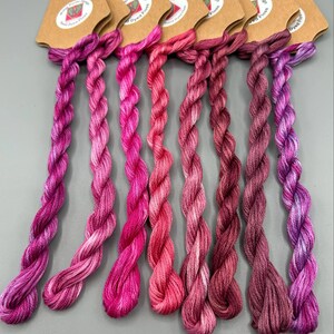May include: Seven skeins of hand-dyed floss in shades of pink and purple. Each skein is attached to a brown paper tag with the text "Winter Design Dyed Floss". The floss is twisted into a rope-like shape.