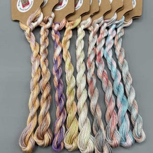 May include: A selection of hand-dyed embroidery floss in pastel shades such as peach, lavender, and blue. Each skein is attached to a brown paper tag. The tag reads "Midge Porter Design Hand Dyed Floss". The text "Light Fantastic': Set 1 of 4" is at the bottom.
